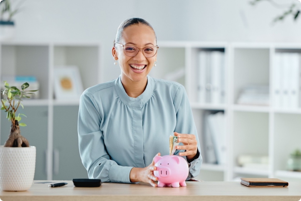 smiling woman in blue shirt puts money into pink piggy bank in office interior setting