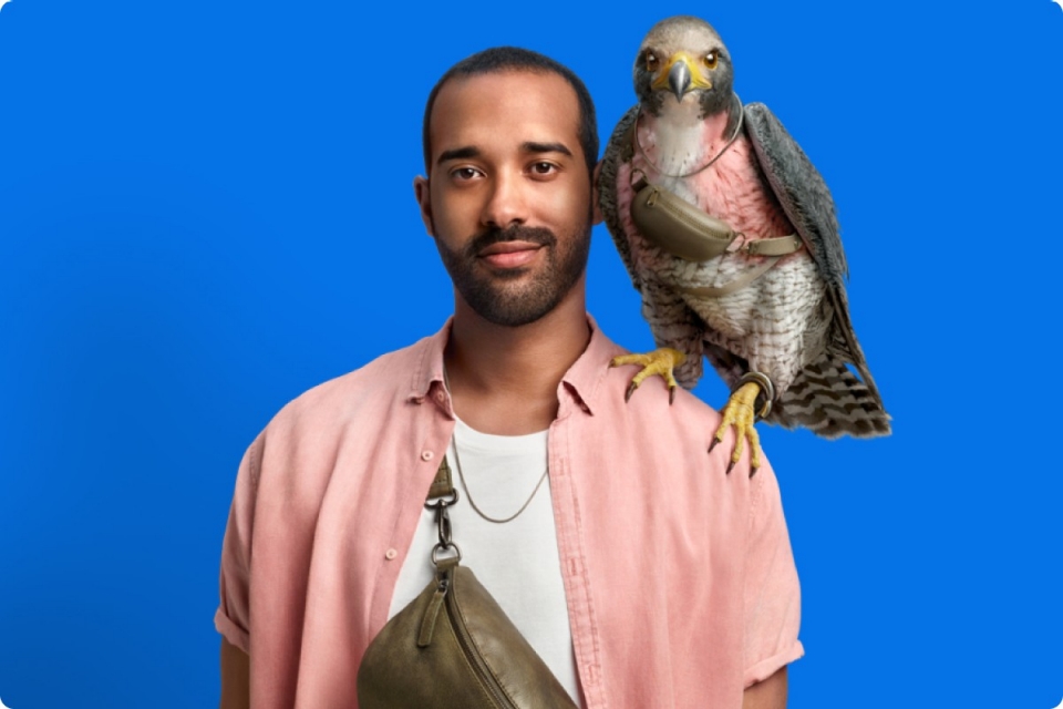 man in salmon coloured overshirt and white undershirt with brown crossbody bag smiles slightly with falcon with similarly coloured feathers around chest perches on his shoulder