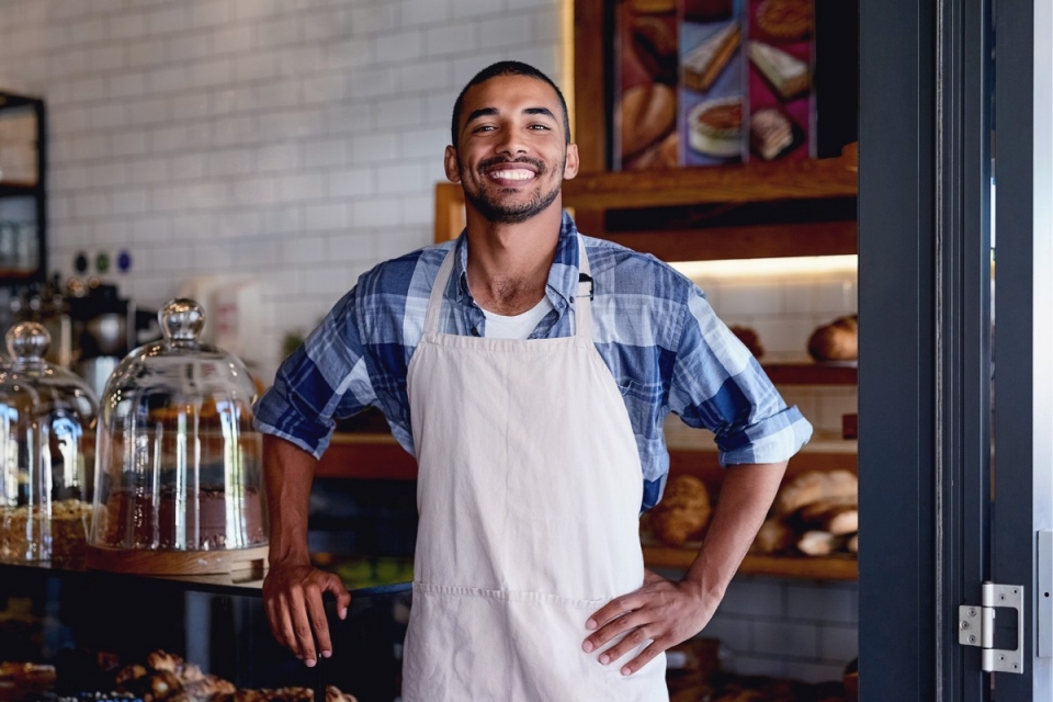 smiling man in blue shirt and white apron leans arm against bakery counter