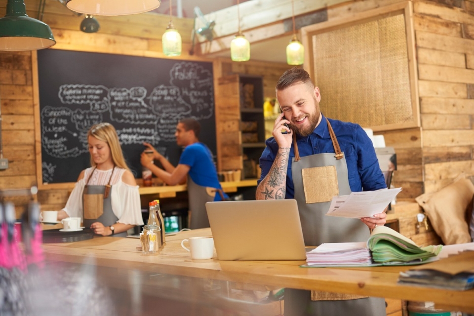 Smiling bearded cafe worker wearing blue shirt and apron on the phone looking at laptop screen with other cafe workers in background
