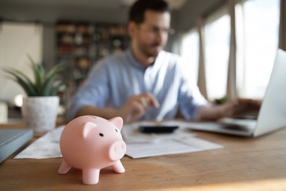 Man working at home on laptop with piggy bank in the foreground 